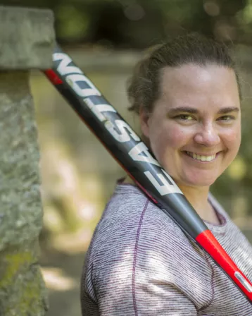 Jen Moore holding a bat and smiling on the whispering bench in the cherry tree glen on campus.