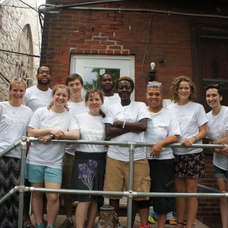 A group of students and Serenity House members, all wearing white T-shirts with the Serenity Soular logo,  pose for a picture on a balcony. 